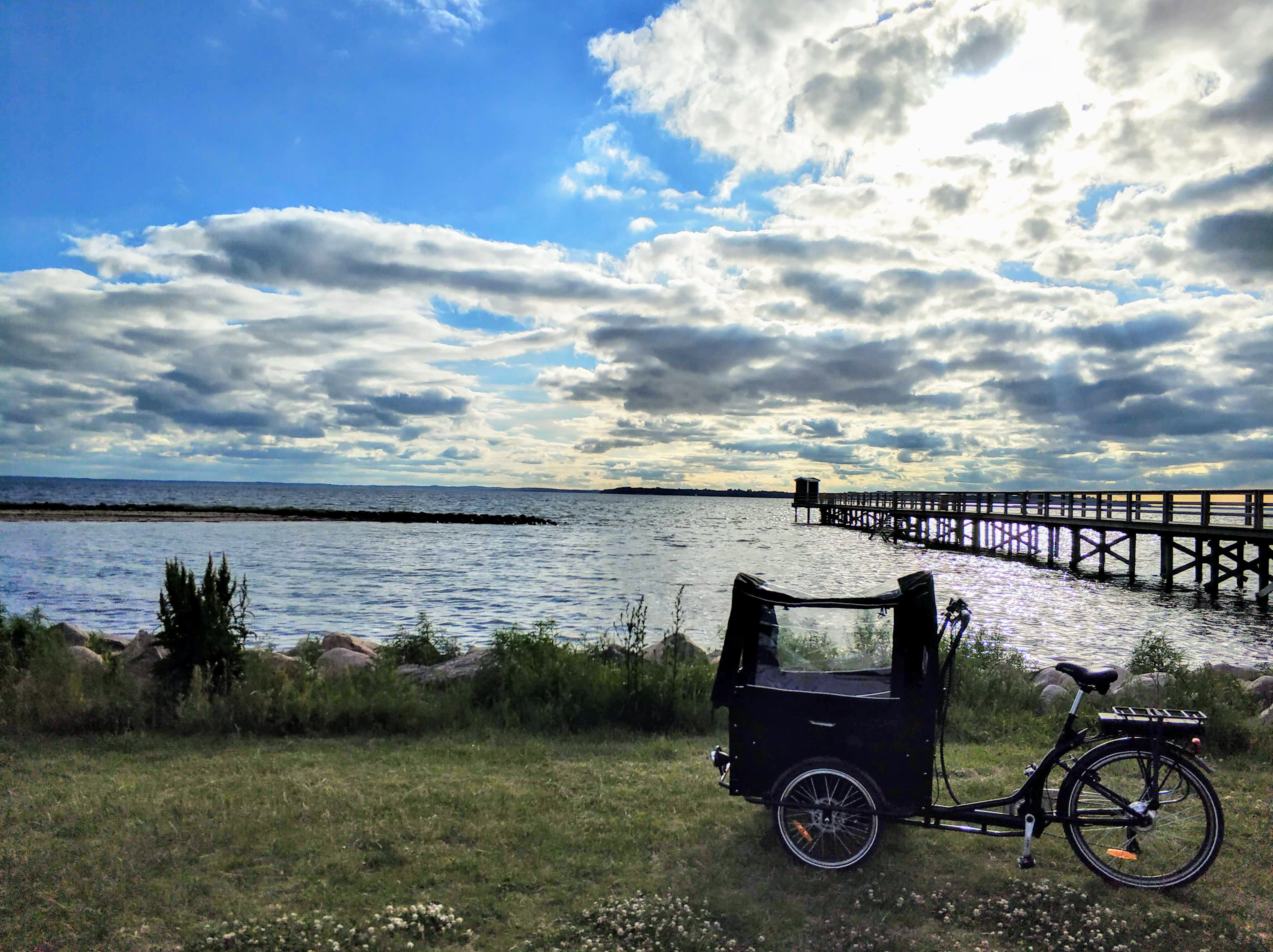 En cykeltaxi står på gräset vid en sjö, med en brygga i bakgrunden och dramatiska moln på himlen