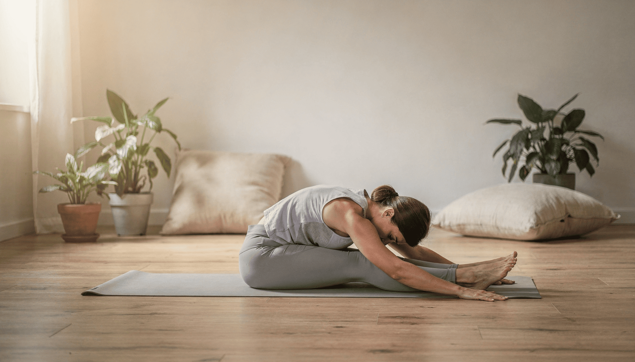 En person utövar stretching i en ljus och lugn inomhusmiljö med växter och kuddar i bakgrunden