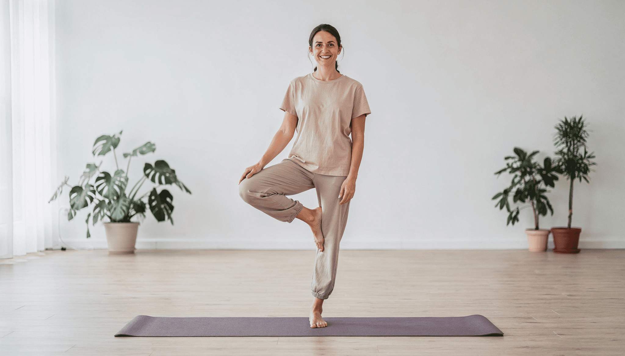 En kvinna står på ett yogamatt och utför en balansövning med leende i en ljus och lugn miljö